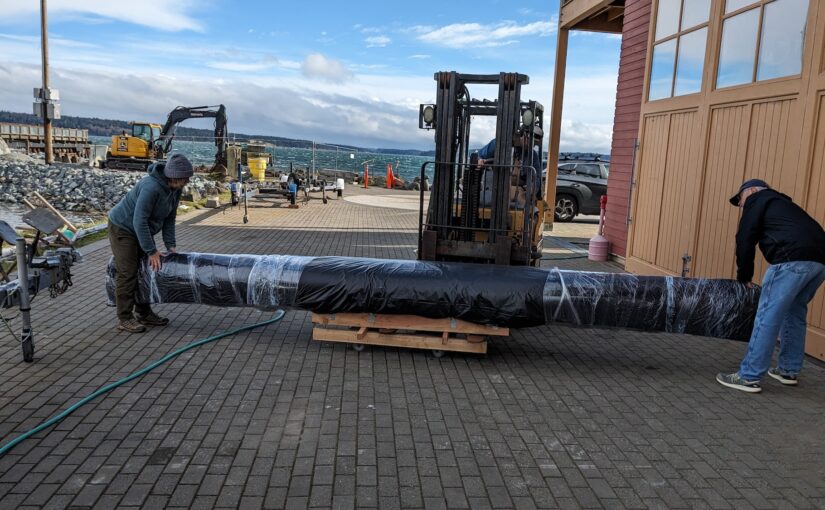 A canoe is wrapped in black plastic. Behind it is a forklift. Janell stands at the bow of the boat, Rich stands at the stern